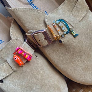 May include: Two tan suede slip-on shoes featuring adjustable straps with metal buckles. The straps are decorated with colorful beaded charms, including an orange fish, a small gold heart, and a miniature blue cowboy boot charm.