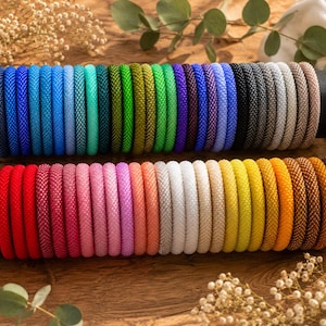 May include: A collection of numerous circular beaded bracelets arranged on two horizontal black cylindrical holders. The bracelets feature a wide spectrum of solid colors including shades of blue, green, purple, red, pink, orange, yellow, white, and black.