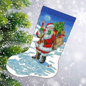 May include: Christmas stocking featuring a detailed illustration of Santa Claus carrying a sack of toys and a drum. The design includes a Christmas tree, a teddy bear, and a snowy landscape. The stocking is white with blue accents.