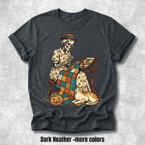 Puede incluir: Una camiseta gris jaspeado oscuro con un gráfico de un esqueleto con sombrero y gafas, sentado en un taburete mientras acolcha una manta de patchwork colorida. Una calabaza naranja y un gato atigrado naranja están en el suelo.