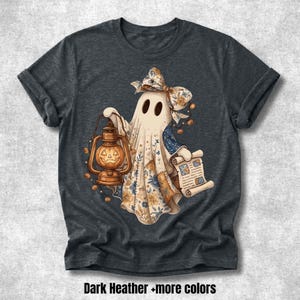 Puede incluir: Una camiseta gris jaspeado oscuro con el gráfico de un fantasma que lleva un lazo con estampado floral, sosteniendo un farol de calabaza iluminado y un pergamino. El fantasma está decorado con pequeñas calabazas naranjas. Texto "Dark Heather +more colors".
