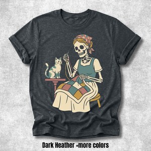 Puede incluir: Una camiseta gris jaspeado oscuro con una ilustración gráfica de un esqueleto con pañuelo en la cabeza cosiendo una colcha de retazos colorida mientras está sentado en un taburete junto a un gato pequeño. El diseño usa tonos apagados de azul, amarillo y rojo.
