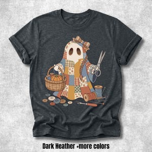 May include: A dark heather gray t-shirt featuring a graphic of a ghost wearing a patchwork quilt, holding a basket with a pumpkin and a pair of scissors. The design includes sewing supplies like spools of thread, buttons, and a seam ripper.