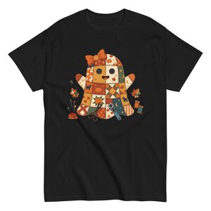 Op de afbeelding: Een zwart t-shirt met korte mouwen en een centrale afbeelding van een geest gemaakt van patchwork-vierkanten in oranje, bruin en crème. De geest draagt een oranje strik en is omringd door naaigerei, waaronder een schaar, een rolsnijder en spelden.