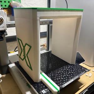 May include: A white and green 3D-printed enclosure with an X-shaped design on the side. The enclosure sits on a black and silver patterned surface, with a green trim along the top and a green stripe on the front edge. The item is on a workbench.