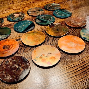 May include: A collection of 15 round coasters with various abstract designs in shades of brown, orange, green, and blue. The coasters are made of a metallic material and have a unique, textured surface.