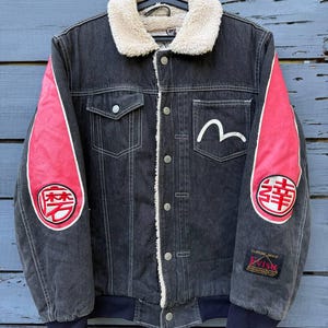 Puede incluir: Una chaqueta de mezclilla negra con cuello de borreguillo color crema, paneles de mangas rosa brillante con parches circulares bordados en rojo y blanco, un logo de gaviota blanco en el bolsillo del pecho y una etiqueta negra con Evisu Custom Made.