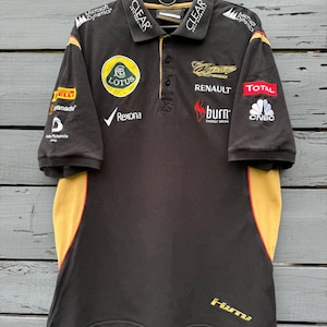 May include: A black polo shirt featuring yellow and red side panels, a collar, and various embroidered sponsor logos including Lotus, Renault, Total, CNBC, Microsoft Dynamics, Clear, Rexona, and Burn. The shirt has short sleeves and a button placket.
