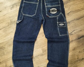 May include: Dark blue denim jeans with multiple pockets and white stitching. The jeans feature the "FUBU" logo on the back pocket and a small logo on the side pocket. The jeans are laid flat on a wooden surface.