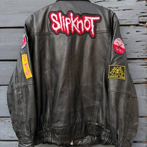 May include: A black leather jacket featuring a large Slipknot patch on the back, a Sex Pistols patch on the right sleeve, and two circular patches on the shoulders. A yellow square patch with a skull and crossbones reads Punk Rock Danger Zone.