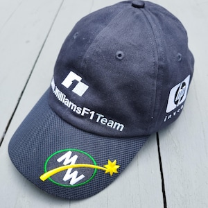 May include: A navy blue baseball cap featuring the white WilliamsF1 Team logo on the front and an HP logo on the side. The brim displays a circular green-bordered emblem with the white letters MW and a yellow shooting star graphic.