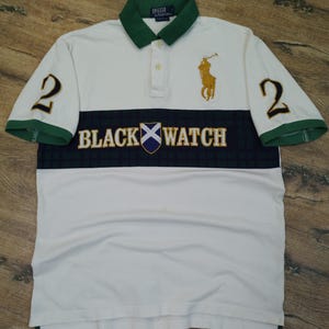 May include: White polo shirt with green collar and sleeve trim. Features the text "BLACK WATCH" across the chest, a gold polo player logo, and the number "2" on both sleeves. The shirt has a button-down collar.
