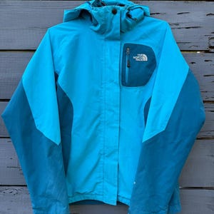 May include: A bright turquoise and dark teal hooded jacket featuring a front zipper closure, a high collar, and a chest pocket with a zipper. The North Face logo is embroidered in white on the upper left chest area of the jacket.
