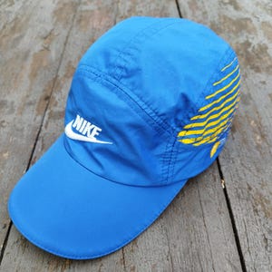 May include: A blue Nike baseball cap with a curved peak. The cap has the Nike logo in white on the front and yellow and gold stripes on the side. It is made of a lightweight material.