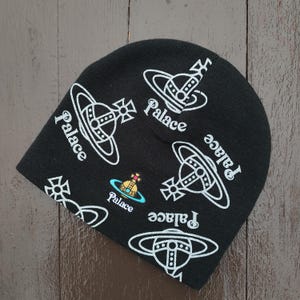 May include: Black beanie hat with white Palace logo and orb graphics. The hat features multiple instances of the Palace text and orb design. A small, colorful orb logo is also present. The hat is made of a soft, knitted material.