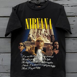 May include: A black short-sleeve t-shirt featuring a graphic print of the band Nirvana. The design includes the yellow Nirvana logo, a photograph of the band members, and the lyrics: I'm not like them but I can pretend, The sun is gone but I have a light, The day is done but I'm having fun, I think I'm dumb or maybe just happy, Think I'm just happy.