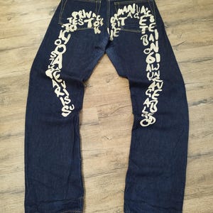 May include: Dark blue denim jeans with white text art on the back of the legs. The text appears to be a series of words and phrases. The jeans have a classic five-pocket design and a brown leather patch.