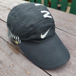 以下が含まれることがあります： 白のブランドロゴが入った濃いグレーのナイキランニングキャップ。キャップにはナイキのスウッシュロゴと「NIKE」の文字が白で入っています。キャップはカーブしたつばと、背面にストライプ模様があります。