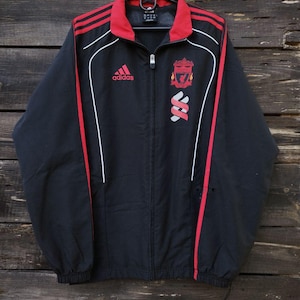 May include: Black Adidas Liverpool jacket with red detailing. Includes a red collar, three red stripes on the sleeves, and white trim. The Liverpool crest and Adidas logo are displayed on the chest.