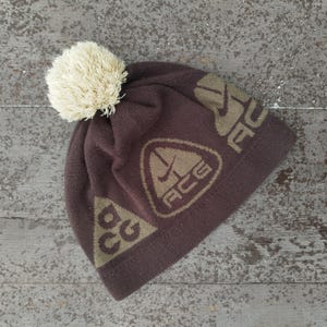 Puede incluir: Un gorro de punto marrón con un pompón color crema. El gorro presenta las letras "ACG" y un logotipo triangular en un color marrón más claro. El gorro está diseñado para el clima frío y actividades al aire libre.