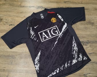 May include: A black short-sleeve soccer jersey featuring a white AIG logo, a Manchester United team crest, and a white Nike swoosh. The shirt has a textured dark gray graphic pattern across the front and a small red accent on the collar.