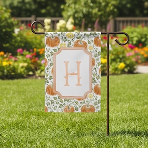 Könnte beinhalten: Eine Gartenflagge mit Kürbis- und Blumenmuster und dem Buchstaben "H" in einem pfirsichfarbenen Rahmen. Die Flagge wird auf einem braunen Metallständer in einem Garten mit Rasen präsentiert.