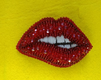 May include: A brooch shaped like a pair of lips, crafted with vibrant red beads and faceted crystals. The design features white thread embroidery to represent teeth, with a distinct black line separating the upper and lower lip sections.