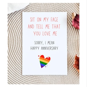 May include: White greeting card with red text that reads "SIT ON MY FACE AND TELL ME THAT YOU LOVE ME. SORRY, I MEAN HAPPY ANNIVERSARY." A rainbow heart graphic is at the bottom. The card is on a textured, neutral surface.