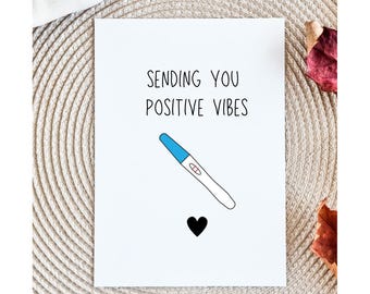 May include: A white card with the text "SENDING YOU POSITIVE VIBES" and a blue and white pregnancy test illustration. A black heart is below the test. The card is on a textured, light-colored surface.