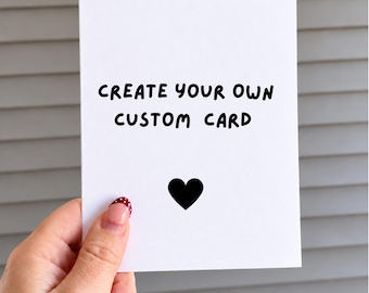 May include: A white rectangular greeting card featuring the black text "CREATE YOUR OWN CUSTOM CARD" in a simple sans-serif font, positioned above a solid black heart icon centered on the lower portion of the card surface.
