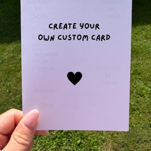 May include: A white rectangular card featuring the black text "CREATE YOUR OWN CUSTOM CARD" centered at the top, with a solid black heart icon positioned below the text. The background of the card contains a faint repeating watermark text.