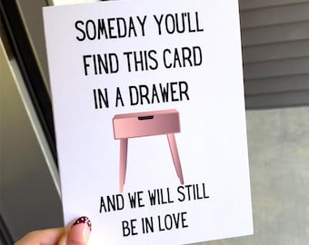 May include: A white greeting card with black text that reads SOMEDAY YOU'LL FIND THIS CARD IN A DRAWER AND WE WILL STILL BE IN LOVE. Centered between the lines of text is a graphic illustration of a pink drawer unit with four legs.