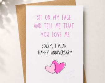 May include: A white greeting card with a brown envelope featuring the pink text SIT ON MY FACE AND TELL ME THAT YOU LOVE ME followed by the black text SORRY, I MEAN HAPPY ANNIVERSARY and two overlapping pink hearts at the bottom center.