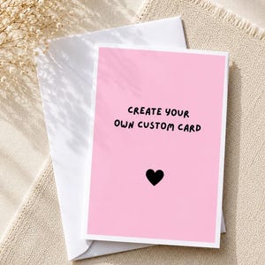 May include: A rectangular greeting card with a solid light pink background and a white border. The text "CREATE YOUR OWN CUSTOM CARD" is printed in black, followed by a small black heart icon. The card is placed on top of a white envelope.