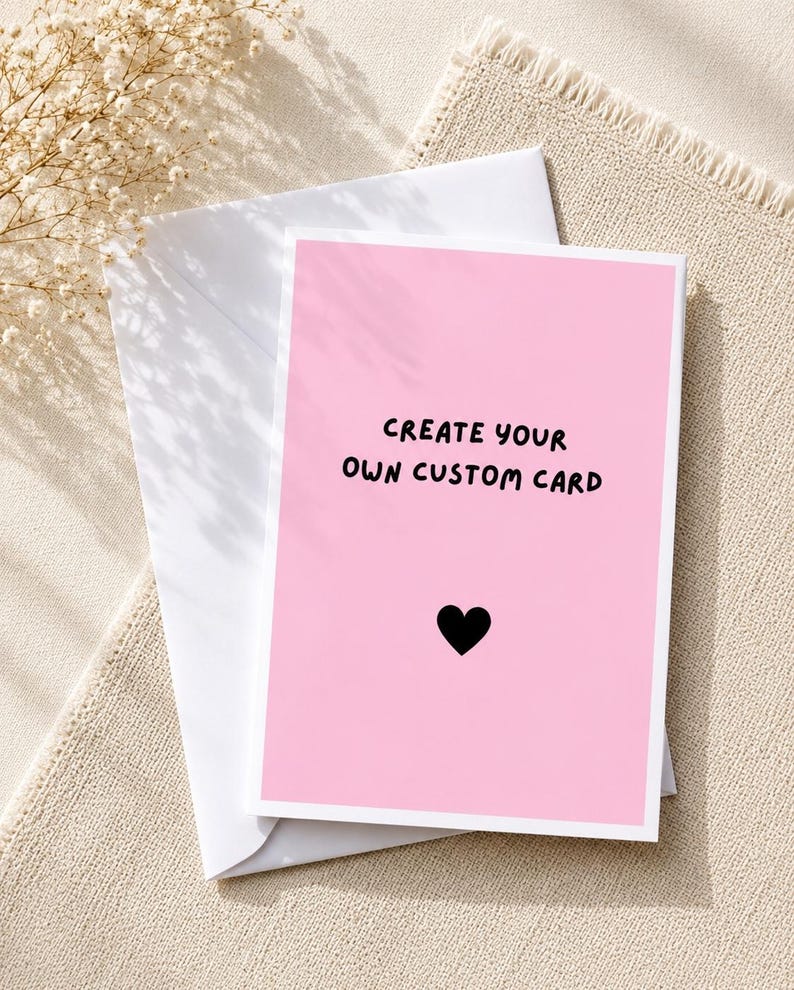May include: A rectangular greeting card with a solid light pink background and a white border. The text "CREATE YOUR OWN CUSTOM CARD" is printed in black, followed by a small black heart icon. The card is placed on top of a white envelope.