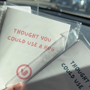 May include: Two white rectangular cards enclosed in clear plastic sleeves. Each card features the text THOUGHT YOU COULD USE A HUG in red or black lettering. One card displays a red circular icon, while the other shows a black circular icon with a heart.