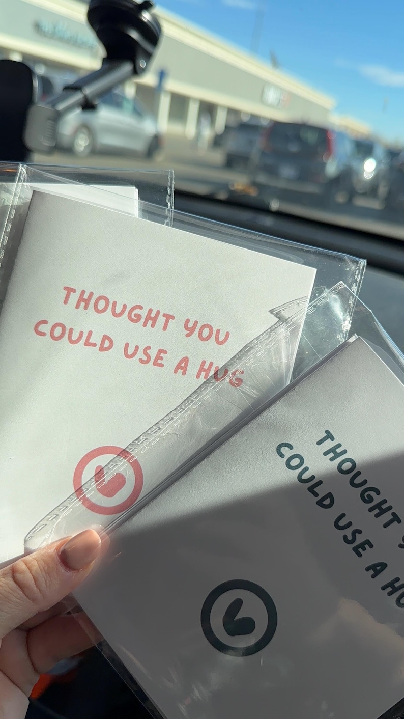 May include: Two white rectangular cards enclosed in clear plastic sleeves. Each card features the text THOUGHT YOU COULD USE A HUG in red or black lettering. One card displays a red circular icon, while the other shows a black circular icon with a heart.