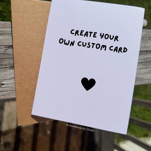 May include: A white greeting card with the black text CREATE YOUR OWN CUSTOM CARD and a small black heart icon below it. The card is paired with a light brown kraft paper envelope. The text Handmade By Chelin appears at the bottom of the card.