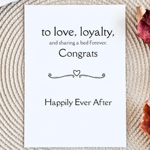 May include: A white greeting card with black text that reads "to love, loyalty, and sharing a bed forever. Congrats. Happily Ever After." The card has a decorative heart and swirl design.