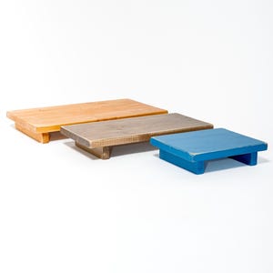 Op de afbeelding: Drie houten display risers in verschillende maten en kleuren. De grootste is van natuurlijk hout, de middelste is grijs en de kleinste is blauw. Elk heeft een rechthoekige bovenkant en een lage, blokvormige basis.