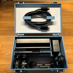 May include: Open, light blue case containing a black game console, charger, headphones, and a controller. A black cable is neatly coiled in the top section. The case is on a wooden surface.