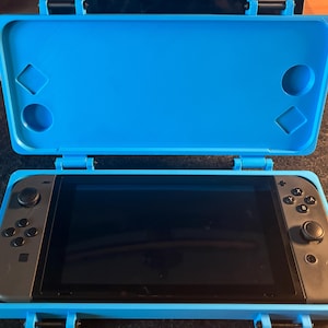 May include: A blue protective case for a Nintendo Switch console. The case is open, revealing the gray console with a black screen. The case has a textured surface and a hinge.