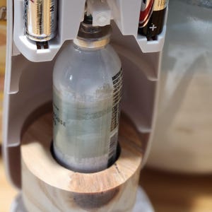 Könnte beinhalten: Ein weißer automatischer Lufterfrischer-Spender mit einem Holzsockel. Der Spender hat eine durchsichtige Plastikflasche mit Lufterfrischer im Inneren. Der Spender wird mit zwei AA-Batterien betrieben. Der Text auf den Batterien lautet "MAX" und "Energizer". Der Text auf dem Spender lautet "OFF 9 18 36".