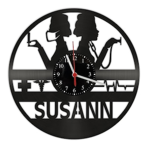 Puede incluir: Reloj de vinilo negro con una esfera blanca y una aguja de hora roja. El reloj presenta una silueta de dos enfermeras con un estetoscopio y un símbolo médico. El nombre "SUSANN" está escrito debajo de la esfera.