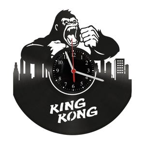 Puede incluir: Reloj de vinilo negro con la silueta de King Kong rugiendo sobre un paisaje urbano. La esfera del reloj tiene números y manecillas blancas, con un segundero rojo. Las palabras "KING KONG" están en texto blanco en la parte inferior.