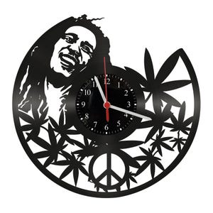 Pode incluir: Relógio de parede preto com a silhueta de um rosto sorridente, rodeado por folhas de cannabis e um símbolo de paz. Os ponteiros do relógio são brancos e vermelhos. O relógio é feito de um disco de vinil.