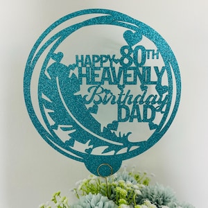 May include: A teal glitter cake topper with the text "Happy 80th Heavenly Birthday Dad" in a circular design. The topper is placed above a bouquet of blue and white flowers, creating a celebratory arrangement.