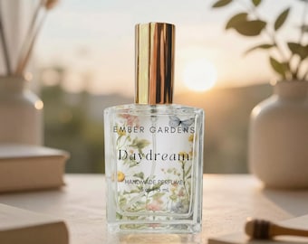 May include: A clear glass perfume bottle with a gold cap, featuring a floral and butterfly print on the front label. The text reads Ember Gardens, Daydream, Handmade Perfume, .5 oz | 15 mL. The bottle has a rectangular shape and contains clear liquid.