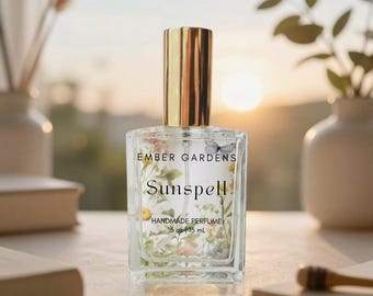 May include: A clear glass perfume bottle with a gold cap, labeled "Ember Gardens Sunspell Handmade Perfume." The bottle features floral and butterfly designs. The perfume is 0.5 oz / 15 mL.