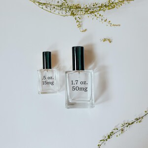 Peut inclure: Deux flacons de parfum en verre transparent avec des bouchons noirs, l'un &eacute;tiquet&eacute; ".5 oz. 15mg" et l'autre "1.7 oz. 50mg". Les flacons rectangulaires sont pos&eacute;s sur une surface blanche, avec des brins verts en arri&egrave;re-plan.